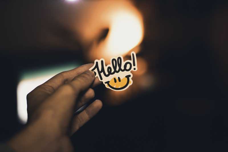 person holding up a "hello" sticker with a smiley face 
